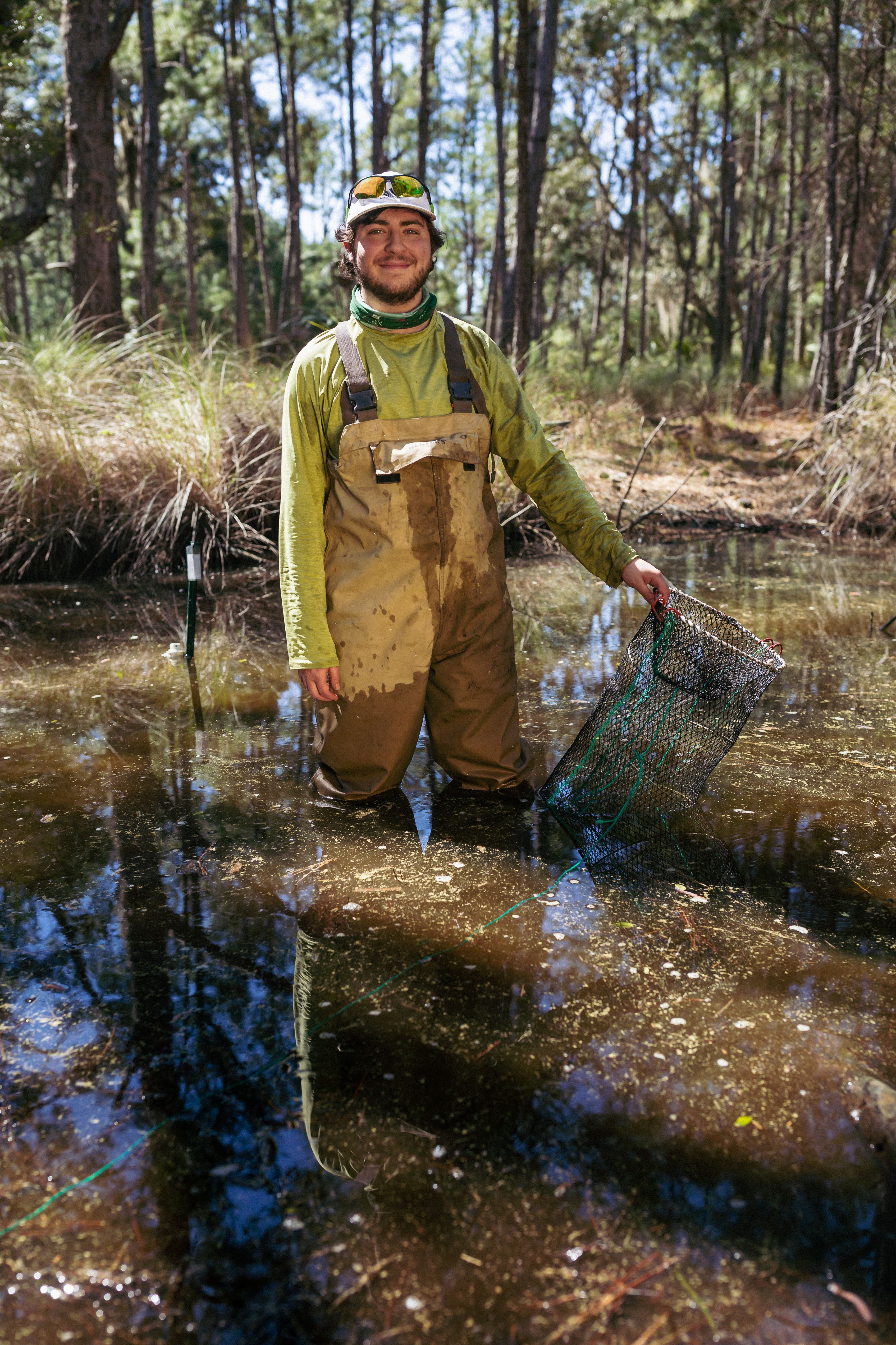 Researcher Zach Gordon standing in a body of water, wearing waiders, and holding an underwater trap