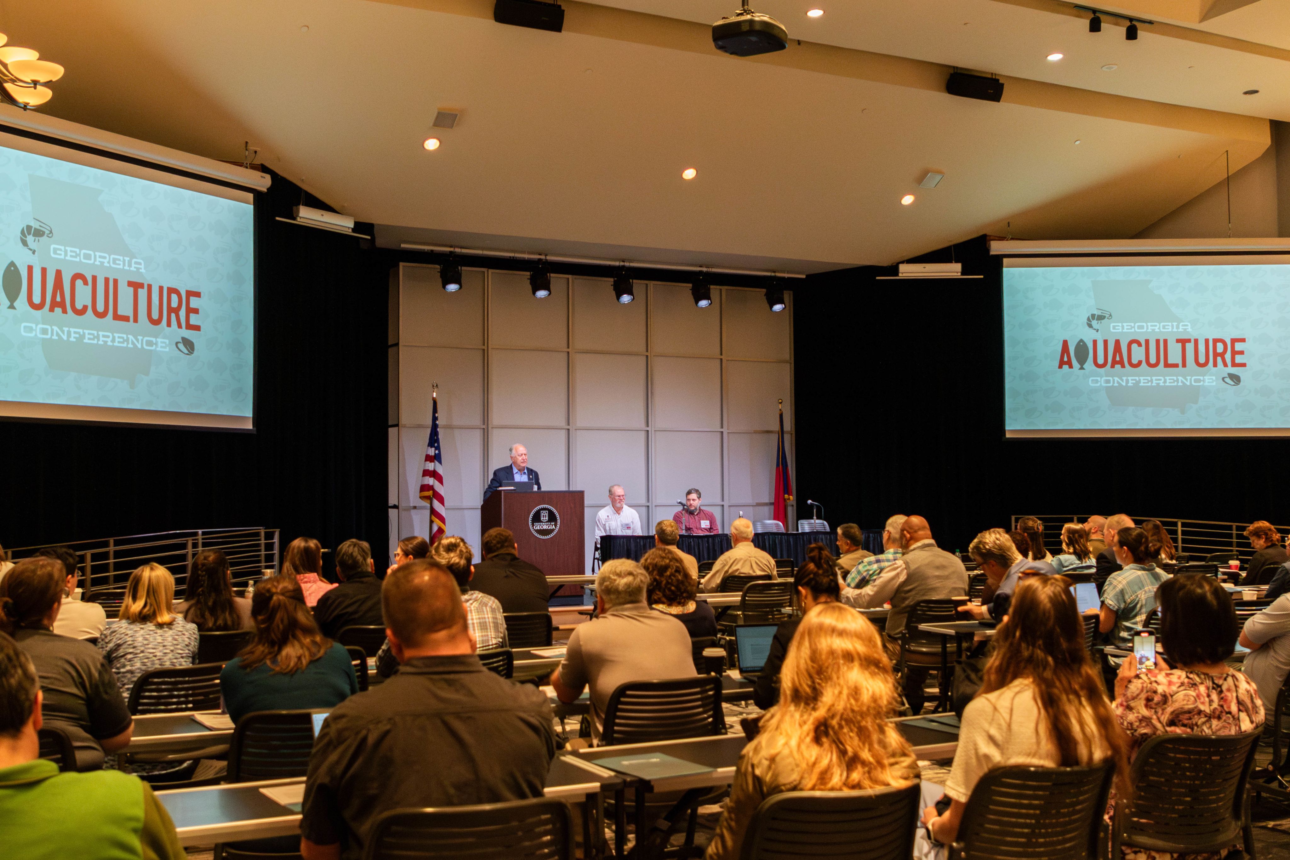 Photo of someone speaking at the Georgia Aquaculture Conference between a podium. An audience is listening intently.
