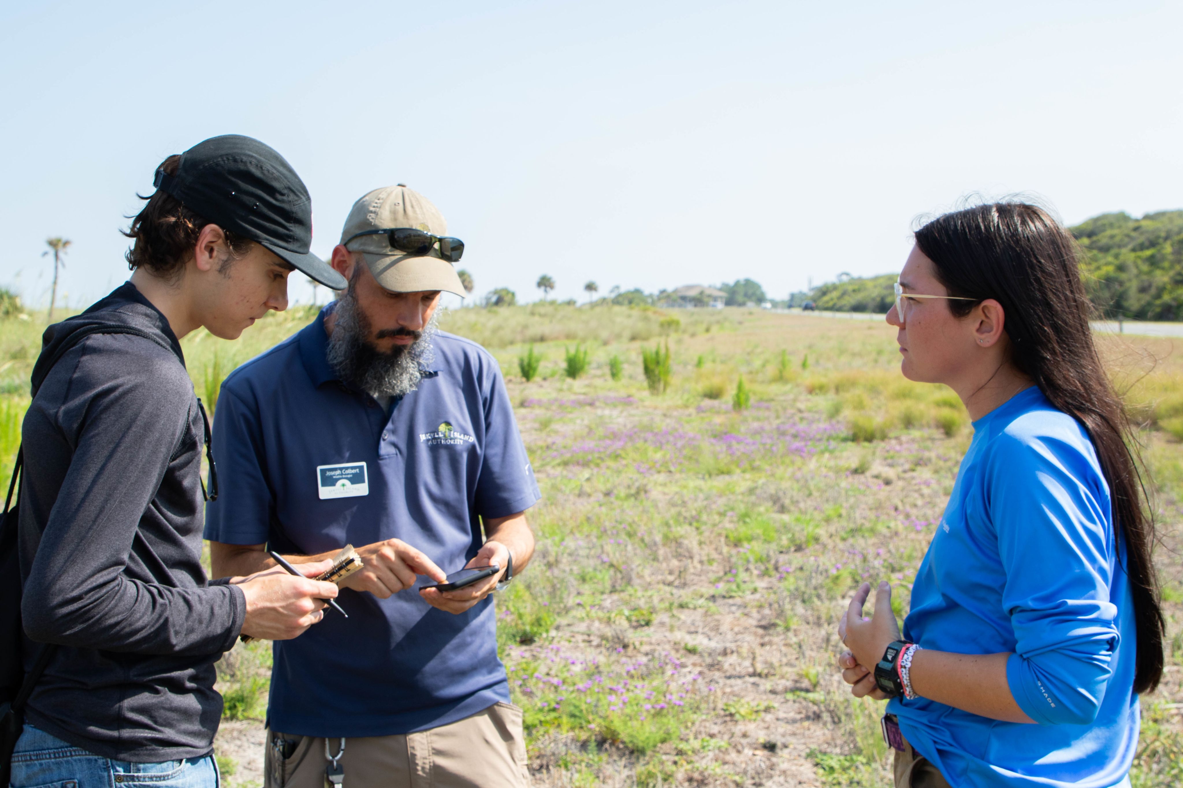 two men and one woman stand outside in a field while they look at a notebook and smart phone